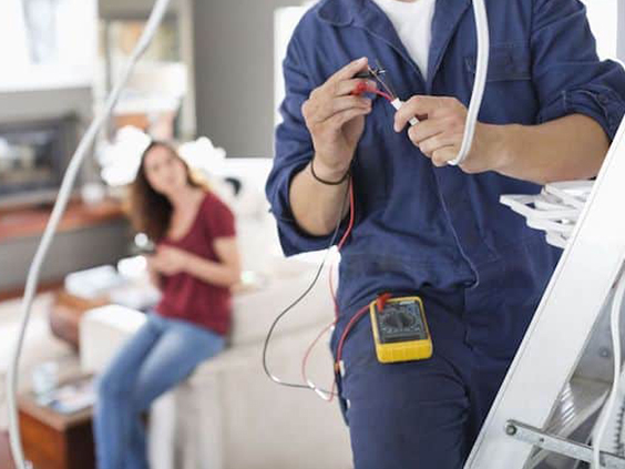Electrician performing wiring work on a ladder in a residential setting, with a female client observing, illustrating hands-on electrical repair services